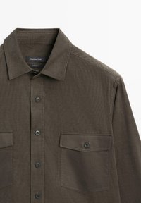 Brązowa koszula sztruksowa z kieszeniami na klapach zapinanych na guziki oraz czarnymi guzikami, kołnierzyk złożony w dół, widoczna metka marki wewnątrz kołnierza.