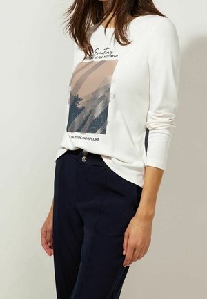 Femme portant une chemise blanche à manches longues avec un motif abstrait et un texte, assortie à un pantalon bleu foncé taille haute avec double boutons.