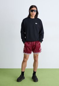 Schwarzes Sweatshirt mit kleinem Logo, rot gemusterte Shorts mit weißen Akzenten und schwarze Slipper, getragen mit übergroßen dunklen Sonnenbrillen.