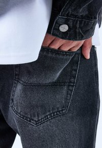 Vaqueros de mezclilla negros con un acabado texturizado, que cuentan con un bolsillo trasero y un puño abotonado en una camisa blanca, mostrando un detalle de atuendo casual.