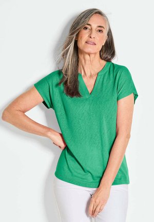Green short-sleeve top with a V-neck design, made of soft fabric. The fabric features a subtle texture, ending at the hips. Paired with white pants.