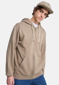 Beige zip-up hoodie gemaakt van zachte stof, met een capuchon, trekkoorden, voorzakken en een geribde onderrand. Gecombineerd met denim shorts.