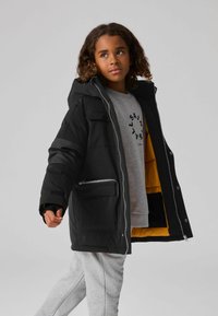 Chaqueta de invierno sintética negra con capucha, que cuenta con un cierre frontal, múltiples bolsillos y un forro interior naranja. Sudadera y pantalones grises visibles.