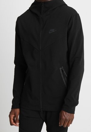 Veste à capuche noire Nike avec fermeture éclair frontale et poches latérales zippées portée par une personne visible du cou jusqu'aux hanches.