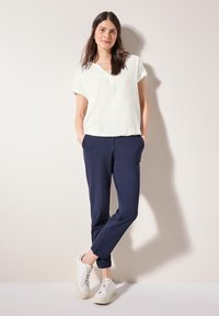 Blouse blanche à manches courtes avec un col en V, associée à un pantalon slim-fit bleu marine et des baskets blanches, se tenant devant un fond neutre.