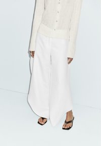 Pantaloni bianchi a gamba larga realizzati in un materiale leggero, abbinati a una camicia beige chiaro con bottoni, caratterizzata da un design a maglia testurizzata.