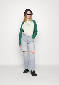 Sweatshirt raglan verde e creme com gráfico "RAGGED", combinado com jeans de cintura alta e desfiados de tom azul claro e sapatos pretos robustos.