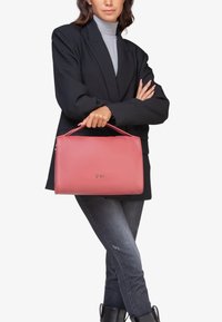 Roze leren handtas met een gestructureerd ontwerp, voorzien van een handvat aan de bovenkant, een gouden logo-accent en een gladde textuur. Draag door een model in casual kleding.