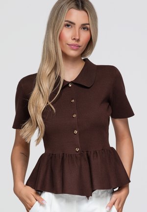 Jeune femme aux longs cheveux blonds portant un haut marron à manches courtes, boutonné, avec un col et un bas peplum, debout les mains dans les poches.