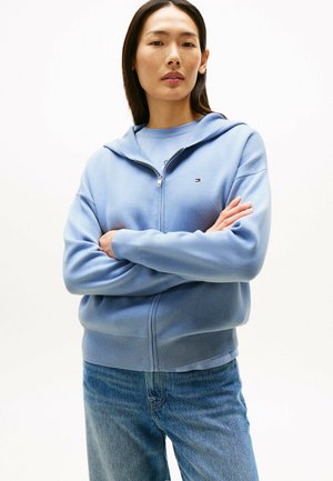 Jasnoniebieska bluza z kapturem na zamek, z małym logo na piersi, w połączeniu ze średnio-pranymi dżinsami. Miękki, teksturowany materiał.