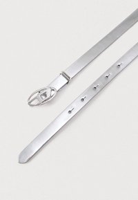 Srebrn usnjen pas z ovalno oblikovano kovinsko zaponko in šestimi enakomerno razporejenimi kovinskimi luknjami na svetlem ozadju.