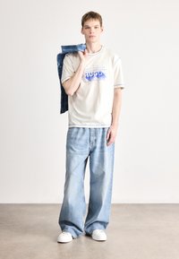 Camiseta bege com gráfico azul e acabamento, combinada com jeans azul claro folgados e ténis brancos. Casaco de ganga pendurado sobre um ombro.