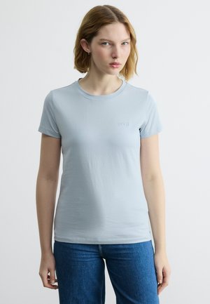 Γυναίκα με κοντά ανοιχτά καστανά μαλλιά, που φοράει ένα ανοιχτό μπλε κοντομάνικο μπλουζάκι και μπλε τζιν, στέκεται μπροστά σε ένα απλό λευκό φόντο.