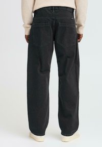 Jeans negre cu picioare largi și talie înaltă, având două buzunare pe spate și o textură ușor estompată. Purtați cu un pulover bej deschis și pantofi albi.
