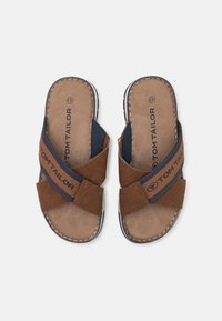 Braune und marineblaue Rutschsandalen mit Kreuzriemen, ausgestattet mit einem strukturierten Obermaterial, weichem Fußbett und einem farblich abgesetzten Etikett. Flache Sohle und kontrastierende Nähte.