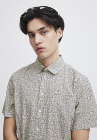 Kortermet skjorte med knapper i lys beige med et blader mønster i blått og mørkere beige. Har krage og hvite knapper.
