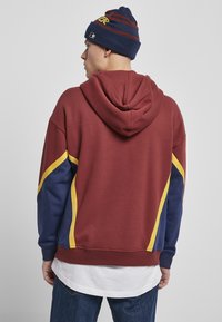 Bordeauxfarbener Kapuzenpullover mit blauen und gelben Akzenten, im entspannten Schnitt und mit strukturiertem Stoff. Getragen mit einer blau gestreiften Mütze.