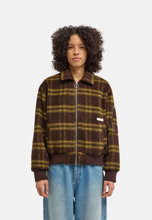 Person mit lockigem Haar, die eine braun-gelb karierte Reißverschlussjacke und lockere hellblaue Jeans trägt, steht vor einem schlichten Hintergrund.