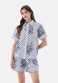 Completo con blusa bianca a maniche corte e pantaloni corti, caratterizzato da motivi floreali e a pois blu navy, chiusura a bottoni e orli ondulati.