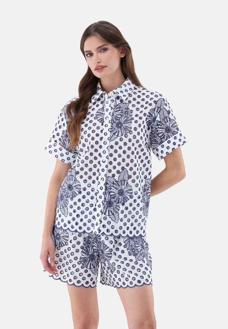 Completo con blusa bianca a maniche corte e pantaloni corti, caratterizzato da motivi floreali e a pois blu navy, chiusura a bottoni e orli ondulati.