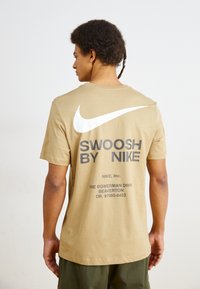Kasta kokvilnas T-krekls ar lielu balto Nike swoosh logotipu un melnu uzrakstu "SWOOSH BY NIKE" uz muguras. Īsas piedurknes, apaļš kakls.