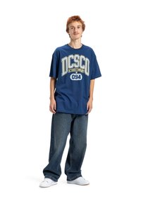 Námornícky modré bavlnené tričko s veľkým bielym a žltým logom "DC SHOE COMPANY 094", skombinované s voľnými tmavými džínsami a bielymi teniskami.