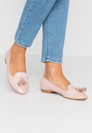 Kvinde iført lyserøde flade loafers med kvaster og lyseblå jeans, stående på en ensfarvet lys gulv.