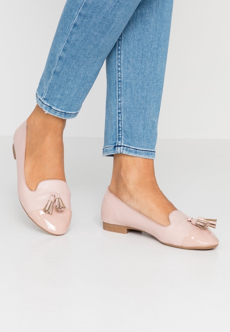 Femme portant des mocassins plats rose clair avec des glands et un jean bleu clair, debout sur un sol clair uni.