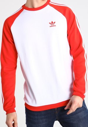 Osoba ubrana w białą bluzę Adidas z czerwonymi rękawami, czerwonym kołnierzem i logo Adidas, w połączeniu z ciemnoniebieskimi dżinsami. Widoczna jest tylko górna część ciała.