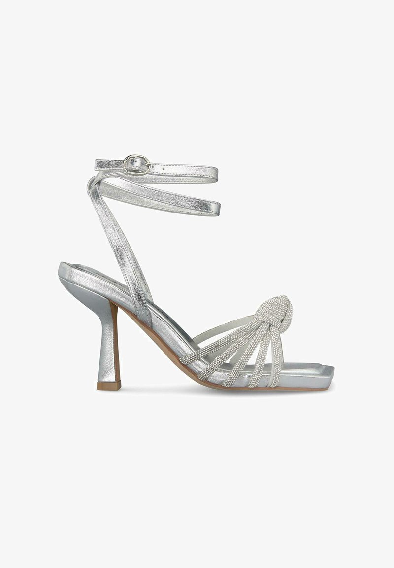 Sandały na srebrnym obcasie z płaskimi paskami i ozdobnym węzłem z przodu. Charakteryzują się eleganckim designem, otwartym noskiem i 7,5 cm obcasem typu stiletto.