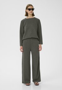 Maglione grigio a maglia con scollatura ampia abbinato a pantaloni grigi a gamba larga. Il modello indossa occhiali da sole neri a forma di gatto e infradito.