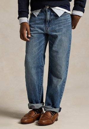 Person iført opsmøgede blå jeans, brune læderloafers, mørk sweater og lagdelte skjorter i en afslappet stående stilling.