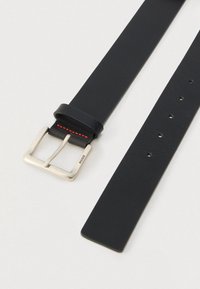 Juodas odinis diržas su sidabrine sagtimi, išgraviruota „HUGO“, ir raudonomis siūlėmis šalia sagties, atidėtas ant baltos paviršiaus.