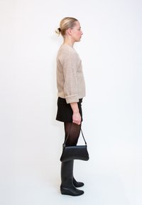 Γυναίκα με μπεζ πουλόβερ, μαύρη φούστα, καλσόν και μπότες κρατώντας μαύρη τσάντα, στέκεται προφίλ σε λευκό φόντο.