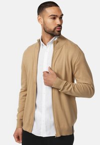 Bežna zip-up cardigan napravljena od mekog materijala, s rebrastim manžetama i visokim ovratnikom, složena preko bijele košulje s dugim rukavima.