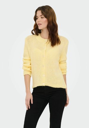 Gelber Strickcardigan mit rundem Ausschnitt, Knopfverschluss und gerippten Bündchen. Weiche Textur und lockerer Schnitt, kombiniert mit schwarzer Hose.