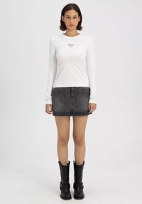 Langärmliges weißes Oberteil mit kleinem Logo, kombiniert mit einem figurnahen schwarzen Jeansrock und knielangen schwarzen Stiefeln mit Schnallenakzenten.