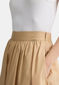 Gonna beige a vita alta realizzata in tessuto morbido, con pieghe e una chiusura in metallo dorato con zip laterale. Texture liscia.