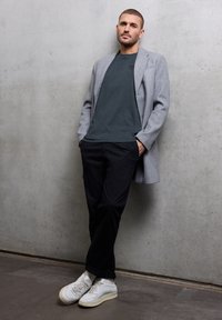 Šedý vlněný kabát přes tmavě zelenou košili, černé kalhoty a bílé tenisky se zelenými akcenty. Hladké textury a pohodlný střih.