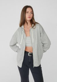 Hellgrau zip-up Kapuzenpullover mit Kordelzug, lockerer Passform und weichem Gefühl. Getragen über einem grauen Oberteil mit kleinen Verzierungen und dunklen Jeans.
