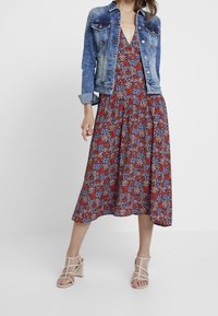 Floral midi dress with a v-neckline in red, blue, and white patterns, layered under a light blue denim jacket; paired with beige block-heeled sandals.