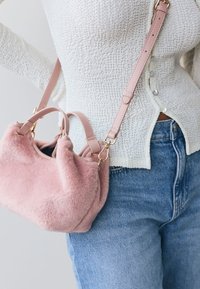 Väike roosa kunstkarva käsikott kahe sangaga ja eemaldatava rihmaga, millel on kuldsed metalldetailid ja pehme, tekstureeritud pind.