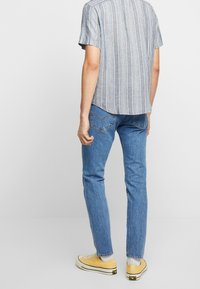 Ljusblå och vit randig kortärmad skjorta i kombination med blå jeans och gula sneakers, sedd bakifrån.