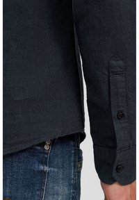 Camisa de manga larga azul oscuro con textura, puños abotonados, bajo ligeramente curvado, combinada con vaqueros de mezclilla con detalles de botones metálicos.