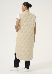 Gilet long beige matelassé avec un col haut, des fentes sur les côtés et une texture lisse. Porté sur un t-shirt blanc et assorti avec un pantalon noir et des baskets blanches.