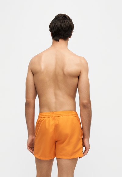 Homme vu de dos portant un short de bain orange vif sur un fond clair uni.