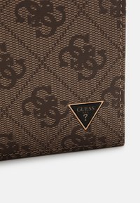 Monedero marrón con un patrón, hecho de material sintético texturizado, que presenta una placa en forma de triángulo con el logo "GUESS" en dorado y negro.