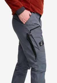 Man som bär grå cargobyxor med flera zippered fickor och en röd fleecejacka, hand vilande i framfickan.