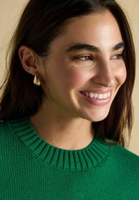 Goldene tropfenförmige Ohrringe mit glatter Oberfläche, vor einem Modell, das einen grünen gerippten Pullover trägt. Glatte Textur und einfaches Design.