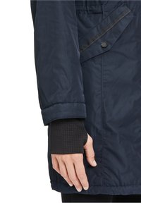 Marineblauer Pufferjacke mit glatter Außenseite, ausgestattet mit einem Kordelzugsaum, Knopfdetails und einem gepufften Ärmel mit einer strukturierten schwarzen Schicht darunter.